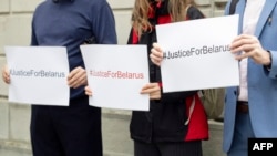 Плякаты з аднолькавым надпісам Justice for Belarus (Справядлівасьць для Беларусі) у руках праваабаронцаў перад слуханьнямі ў крымінальнай справе ва ўнівэрсальнай юрысдыкцыі